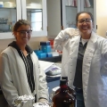 Two people in lab coats and safety goggles smiling in a laboratory.