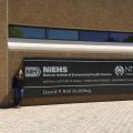 Woman standing next to a sign for the NIH National Institute of Environmental Health Sciences and National Toxicology Program on a brown, textured building wall.