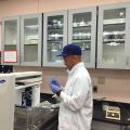 Man in lab coat and cap working with lab equipment in a laboratory with gray storage cabinets and a handwritten sign.