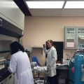 Laboratory scene with two individuals in white lab coats, laboratory equipment, storage cabinets, and large gas cylinders.