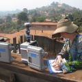 Person in a straw hat writing on a clipboard next to two grey devices, with a densely populated hilly area in the background.