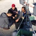 Three people crouched around an air monitor setup on a concrete surface under bright sunlight.