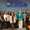 A group of twelve people are standing in front of a blue wall with a sign reading "COUNTY OF SAN DIEGO HHSA HEALTH AND HUMAN SERVICES AGENCY."