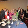 A group of eleven people, six women and five men, are gathered around a formally set dining table at an indoor event.