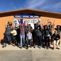 Group of people jumping in front of an orange building with signs.