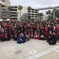 Group of university graduates in caps and gowns posing for a photo outdoors near a parking structure.