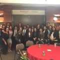 Group photo of 23 people in business attire in front of the Stephen L. Weber Alumni Board Room. Tables with red tablecloths and beverage cans are in the foreground.