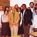 A diverse group of people standing together in a classroom setting, smiling at the camera. A chalkboard in the background has the words "SAFE ALL STUDENTS" written on it.