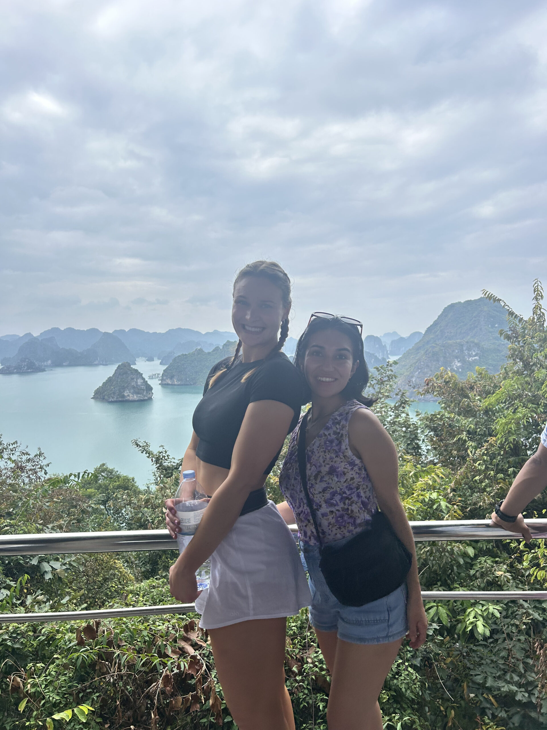 Two people posing in front of a scenic bay with rocky islands and cloudy skies.