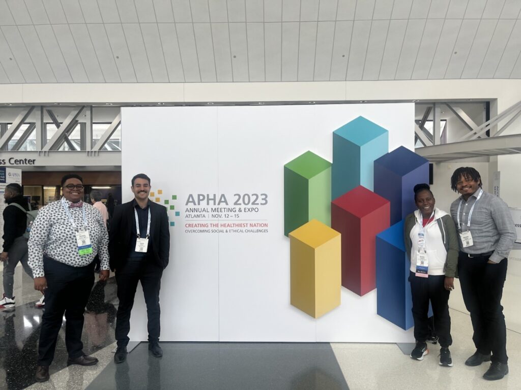 Michelle Caesar, Jacob Carson, Doreen Tuhebwe, and Giovanni Appolon pose at the entrance to the APHA 2023 Annual Meeting & Expo in Atlanta. 